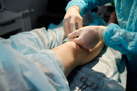 Gloved hands of professional surgeon holding syringe with anaesthesia injection by knee of young female patient on operation tableの写真素材