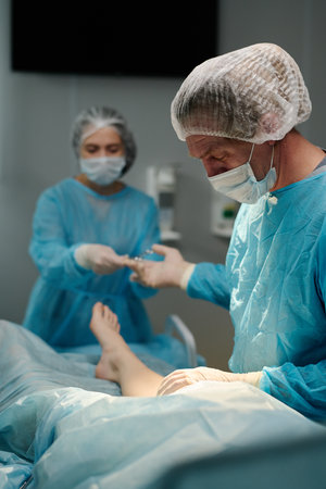 Mature surgeon taking medical tool from hand of female assistant in medical scrubs, gloves, cap and mask during operationの写真素材