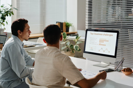 Two young male managers looking through online statistic data on screen of desktop computer while sitting by workplace in officeの写真素材
