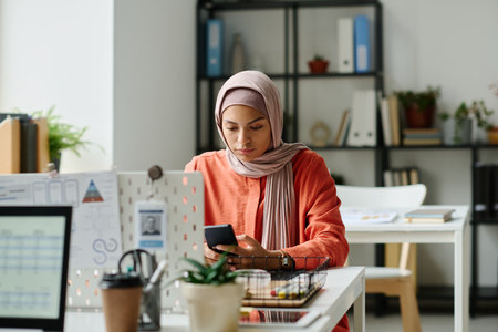 Young Muslim female office worker in hijab sitting by workplace in coworking space and scrolling through posts in smartphoneの写真素材