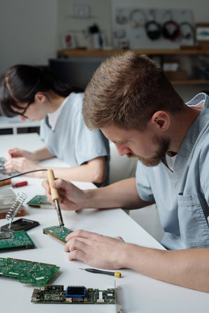 Young man with electric handtool bending over desk and motherboard while repairing detail of computer processor against colleagueの写真素材