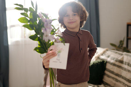 Cheerful boy looking at camera with smile while passing handmade postcard and bunch of flowers to his mother on holidayの写真素材