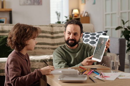 Bearded father with tablet sitting by small table and showing his son example of postcard for mother day on screen of mobile gadgetの写真素材