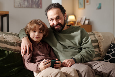 Happy young affectionate man embracing his cute little son with mobile phone while both sitting on...