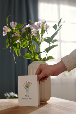 Hand of unrecognizable woman putting handmade greeting card for her mother against white vase with bouquet of fresh flowersの写真素材