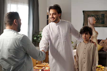 Medium shot of young Muslim man and his son wearing thobes greeting male relative on Eid al-Fitrの写真素材