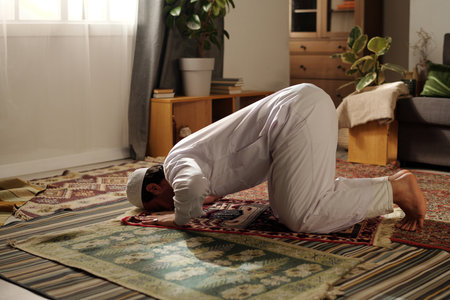 Young Muslim man wearing white clothes doing sajdah while practicing namaz on prayer rug in living room at homeの写真素材