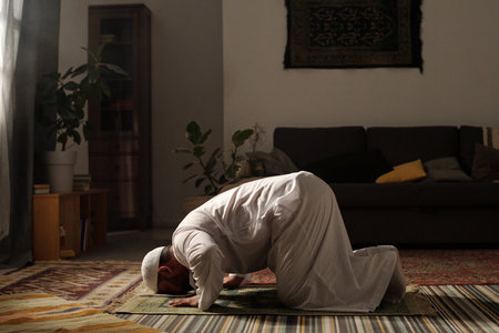 Long shot of mature bearded Muslim man wearing white thobe doing sajdah on prayer rug in living room at home, copy spaceの写真素材