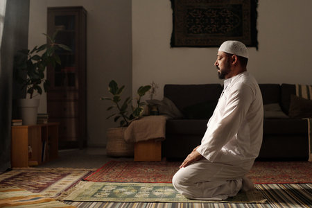 Side view shot of mature Muslim man wearing white clothes sitting on prayer rug in living room doing namazの写真素材