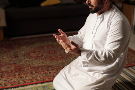 High angle view shot of bearded Muslim man in white clothes sitting on rug praying salah, copy space