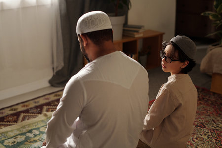 Muslim father and son wearing takiyahs sitting on rugs on floor in living room praying salahの写真素材