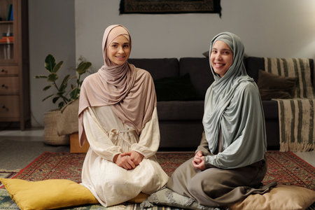 Two happy young Muslim women wearing hijabs sitting on floor in living room smiling at camera, medium long portraitの写真素材