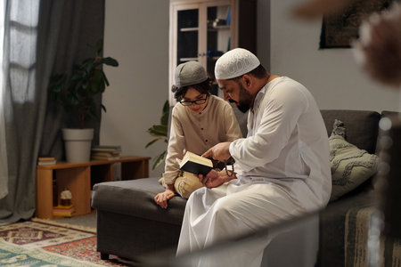 Modern bearded Muslim man wearing kandora sitting on sofa in living room explaining Quran surah to his son, copy spaceの写真素材