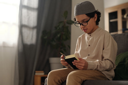 Medium shot of Muslim boy wearing eyeglasses sitting on couch reading holy Quran book, copy spaceの写真素材