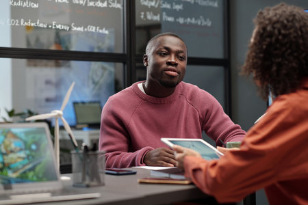 Young African American businessman in casualwear looking at female colleague with tablet and...