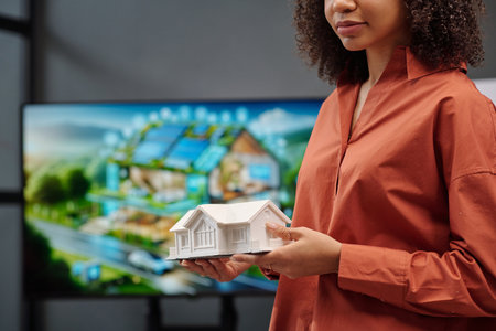Serious female architect in brown shirt holding model of house while standing against screen with visual template of her projectの写真素材