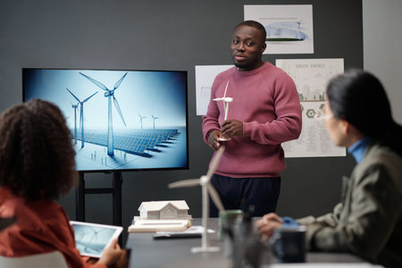 Young businessman with model of windmill in hands looking at female colleague while standing by interactive board during presentationの写真素材