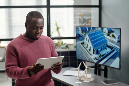 African American male employee in casualwear networking or scrolling through project points against his workplace with interactive boardの写真素材