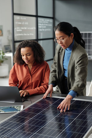 Young African American woman typing on laptop keyboard while sitting next to Asian colleague making presentation of solar panel modelの写真素材