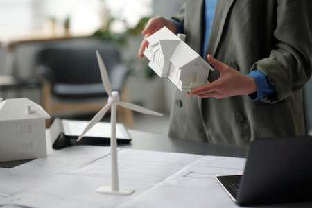 Cropped shot of young unrecognizable female architect in grey suit holding model of house over workplace with unrolled blueprintの写真素材
