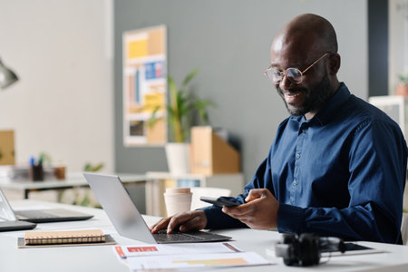 Medium shot of cheerful African American manager using smartphone and typing on laptop at work in modern office, copy spaceの写真素材