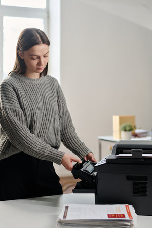 Vertical medium long shot of young Caucasian woman working in modern office taking ink cartridge out...