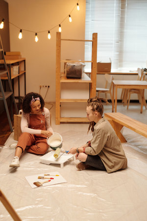 Two young women sitting on the floor of cafe while one of them pouring paint in tray while preparing it for renovation of interiorの写真素材