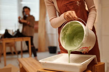 Cropped shot of young woman in overalls bending over bench and pouring green paint in plastic tray from bucket during cafe refitの写真素材