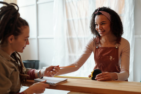 Young smiling female worker or designer in overalls taking measures of bench and looking at her...