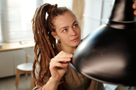 Young female owner of cafe or restaurant with dreadlocks looking at new lamp while hanging it over table during renovation worksの写真素材