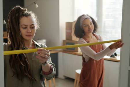 Female worker or designer with dreadlocks and her colleague holding measuring tape on both sides of...