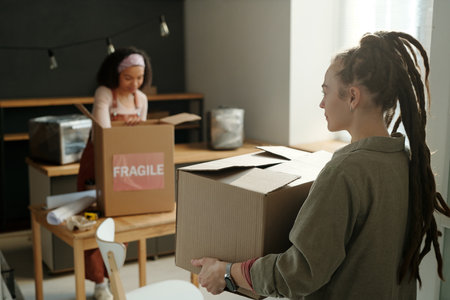Young businesswoman with dreadlocks carrying packed cardboard box while moving towards her colleague...