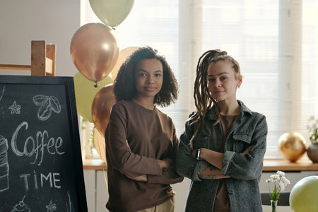 Two young intercultural female entrepreneurs in casualwear looking at camera while standing in...