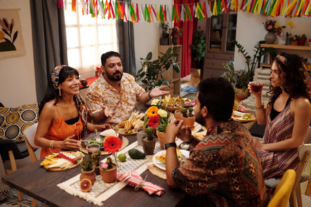 Group of young friends having chat by served festive table while enjoying dinner with homemade food in living room decorated with pennantsの写真素材