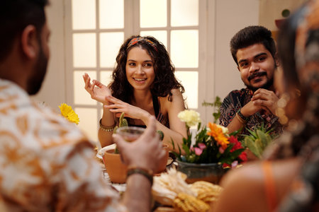 Young smiling brunette woman looking at one of friends during chat by festive table at home party...