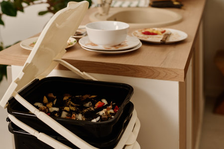Plastic bucket or trash bin with open lid containing leftovers of organic food standing by kitchen...
