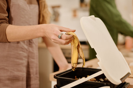 Hand of young woman with banana peel throwing it in trash bin or bucket with open lid while standing in front of cameraの写真素材