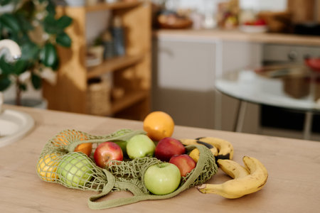 Variety of fruits in shopping bag lying on kitchen counter or wooden table standing against home interior with green domestic plantsの写真素材