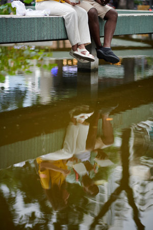 Reflection on water surface of young travelers sitting on architectural construction in urban...