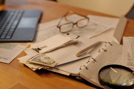 Pen, stack of receipts and envelope with dollars lying on pages of open notebook next to magnifying...