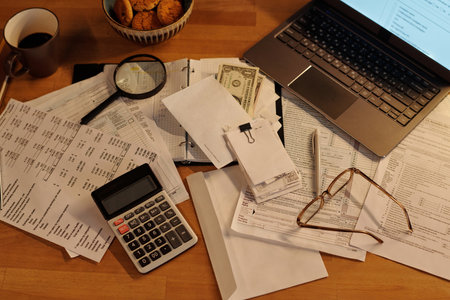 High angle of calculator, envelope with dollar banknotes, magnifying glass, stack of receipts, pen, eyeglasses, financial bills and laptop on tableの写真素材