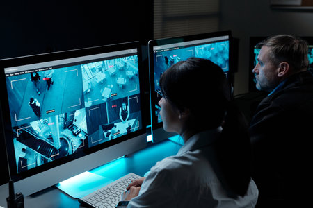 Two intercultural security guards in uniform sitting by workplace in front of computer monitors and...