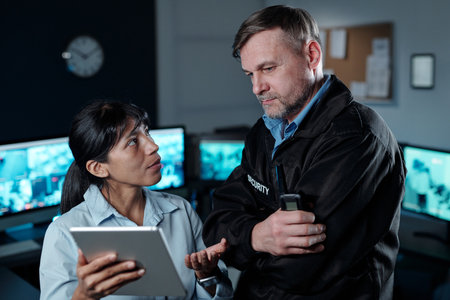 Young Hispanic female security guard with tablet looking at mature male colleague during...