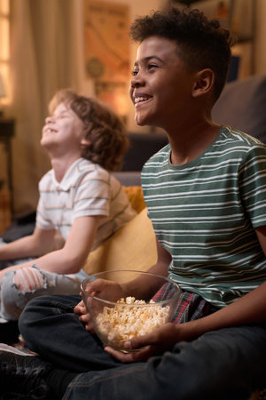 Vertical shot of two multiethnic schoolboys laughing and watching TV while sitting on floor in cozy living room, enjoying time together with popcorn and a relaxed atmosphereの写真素材