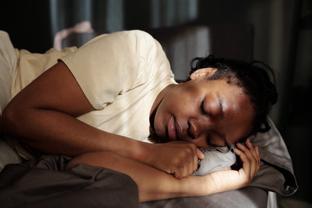 Young depressed woman in t-shirt keeping her head on pillow while lying on bed in front of camera at night and trying to sleepの写真素材