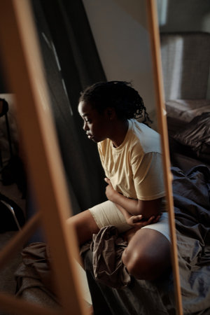 Reflection in mirror of young desperate woman sitting on bed and concentrating on thoughts caused by post traumatic syndromeの写真素材