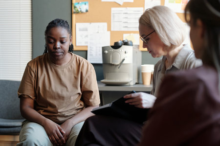 Young depressed African American woman sitting in front of psychologist making notes in notepad...