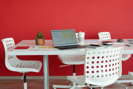 Modern office workspace with red wall in background, featuring laptop, coffee cup, and other office...