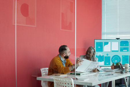 Two colleagues, Caucasian man and African American woman, discussing documents in modern office with red walls and presentation screen in backgroundの写真素材