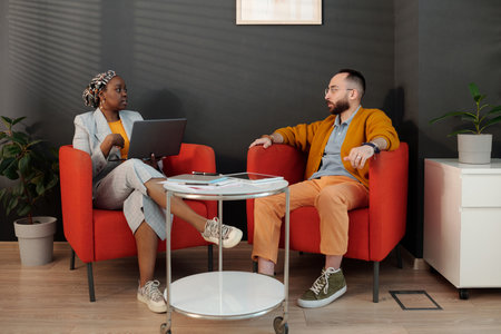 Two colleagues having discussion in modern office lounge. African American woman using laptop while Caucasian man listening attentively, both seated on red chairs around small tableの写真素材